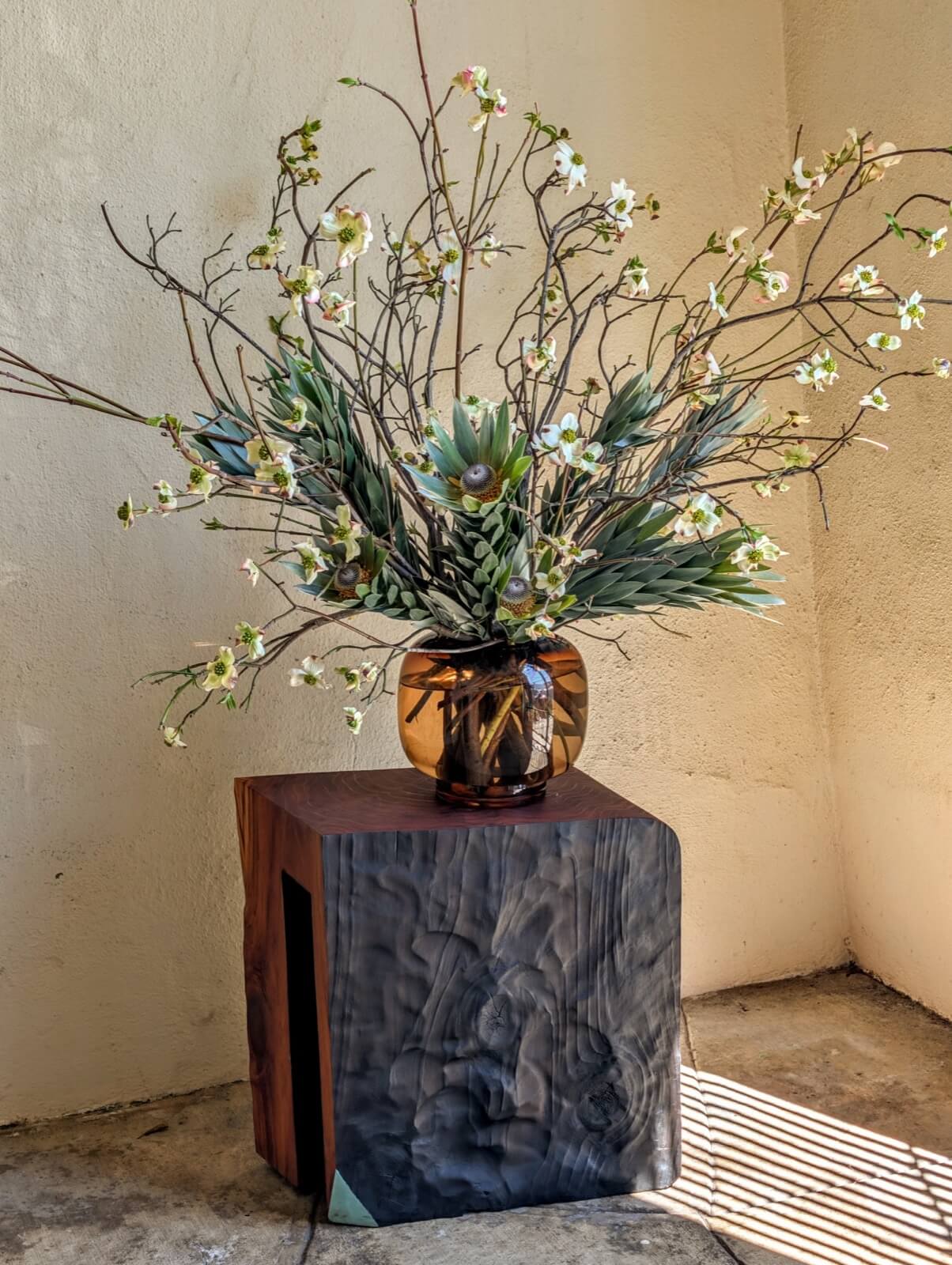 Branching arrangement of dogwood and grasses in an amber glass vase on a charred wood block