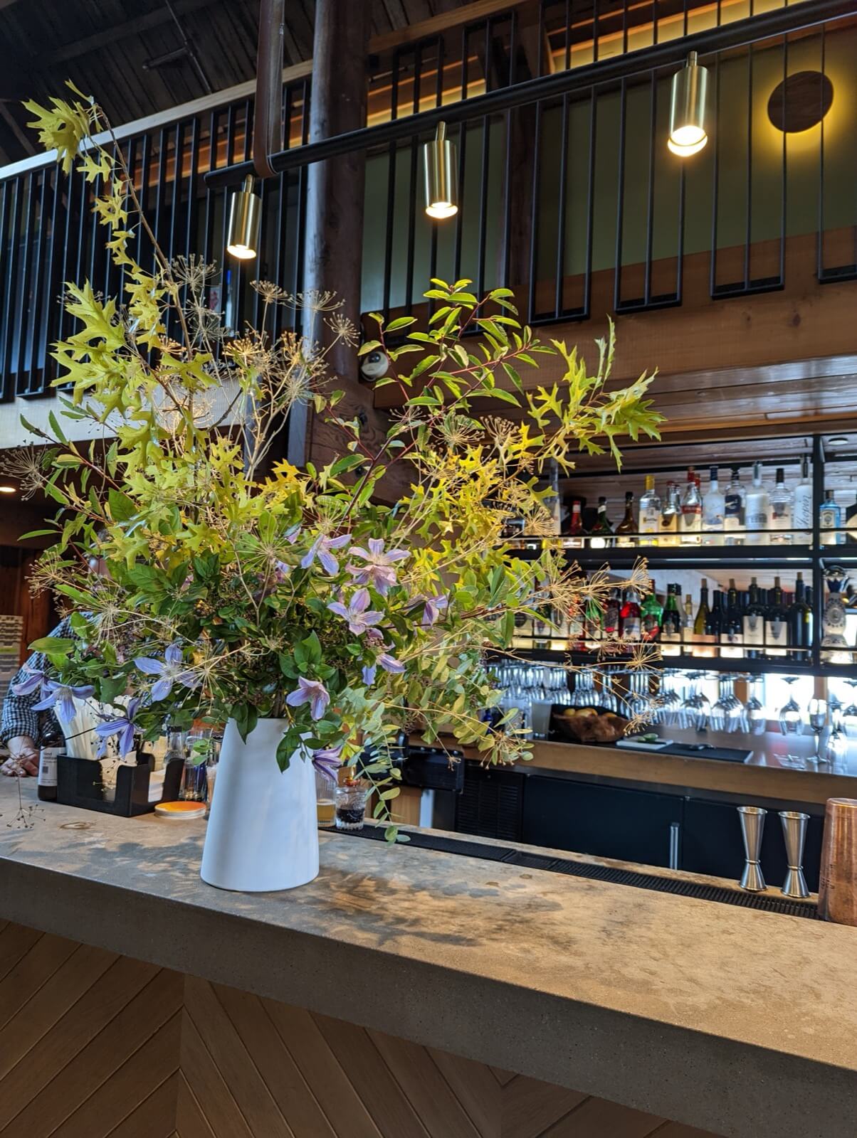 Loose greenery and clematis in a white pitcher on a restaurant bar