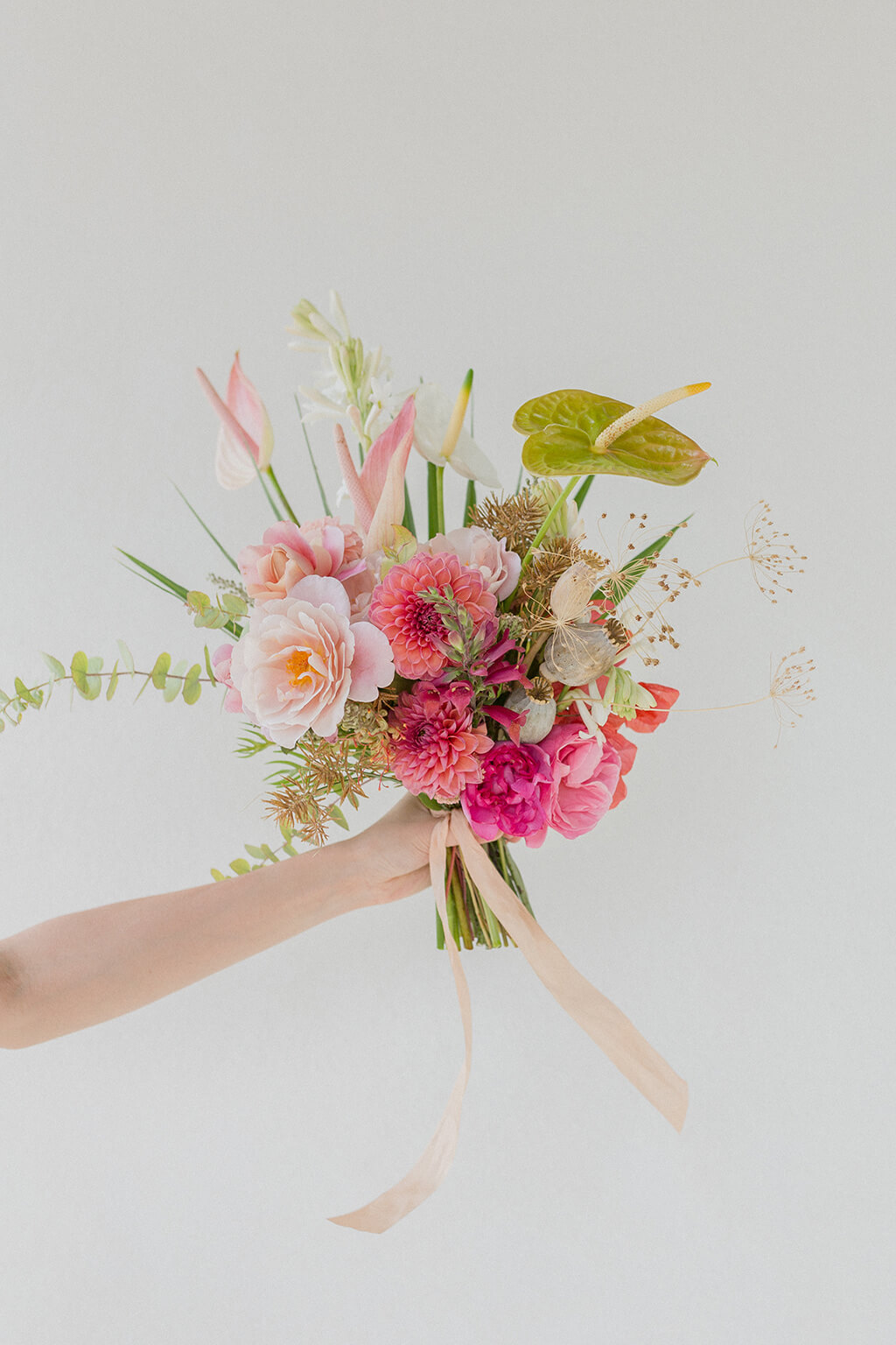 Hand holding a bouquet of pink dahlias, anthurium, and tuberose with trailing ribbon