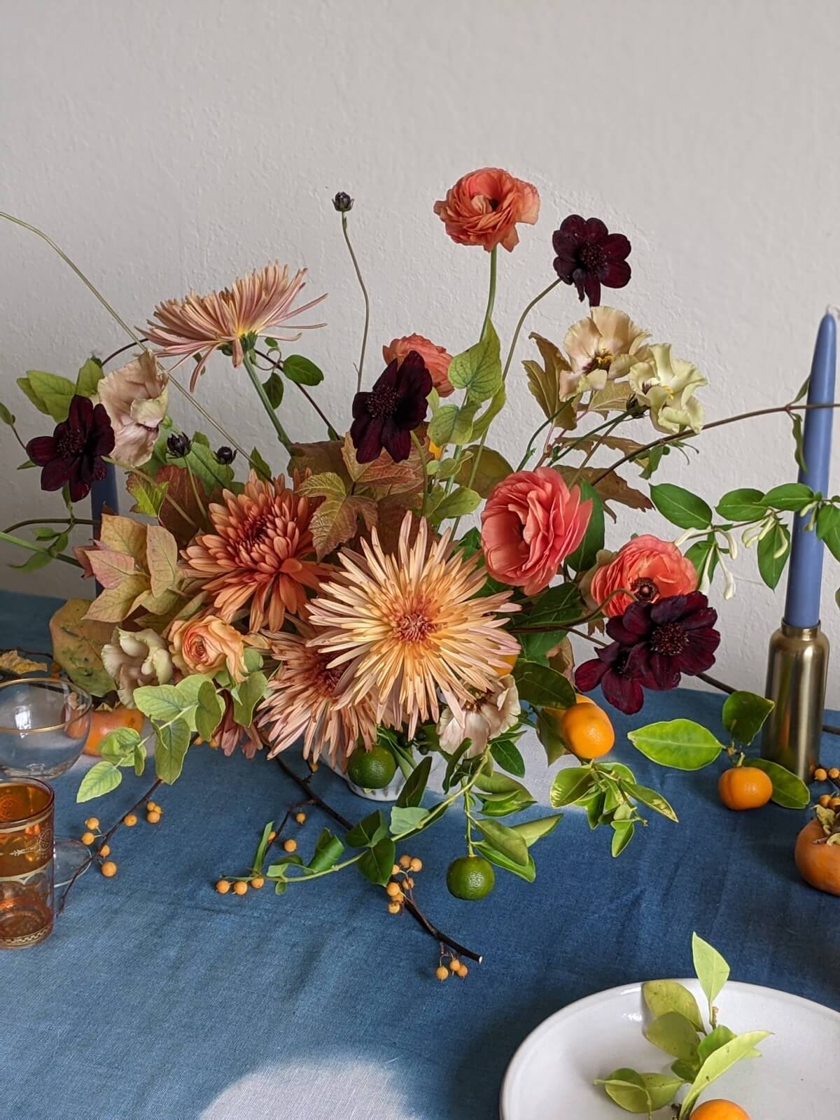 Peach chrysanthemums, ranunculus, and chocolate cosmos with kumquats on a blue linen tablecloth