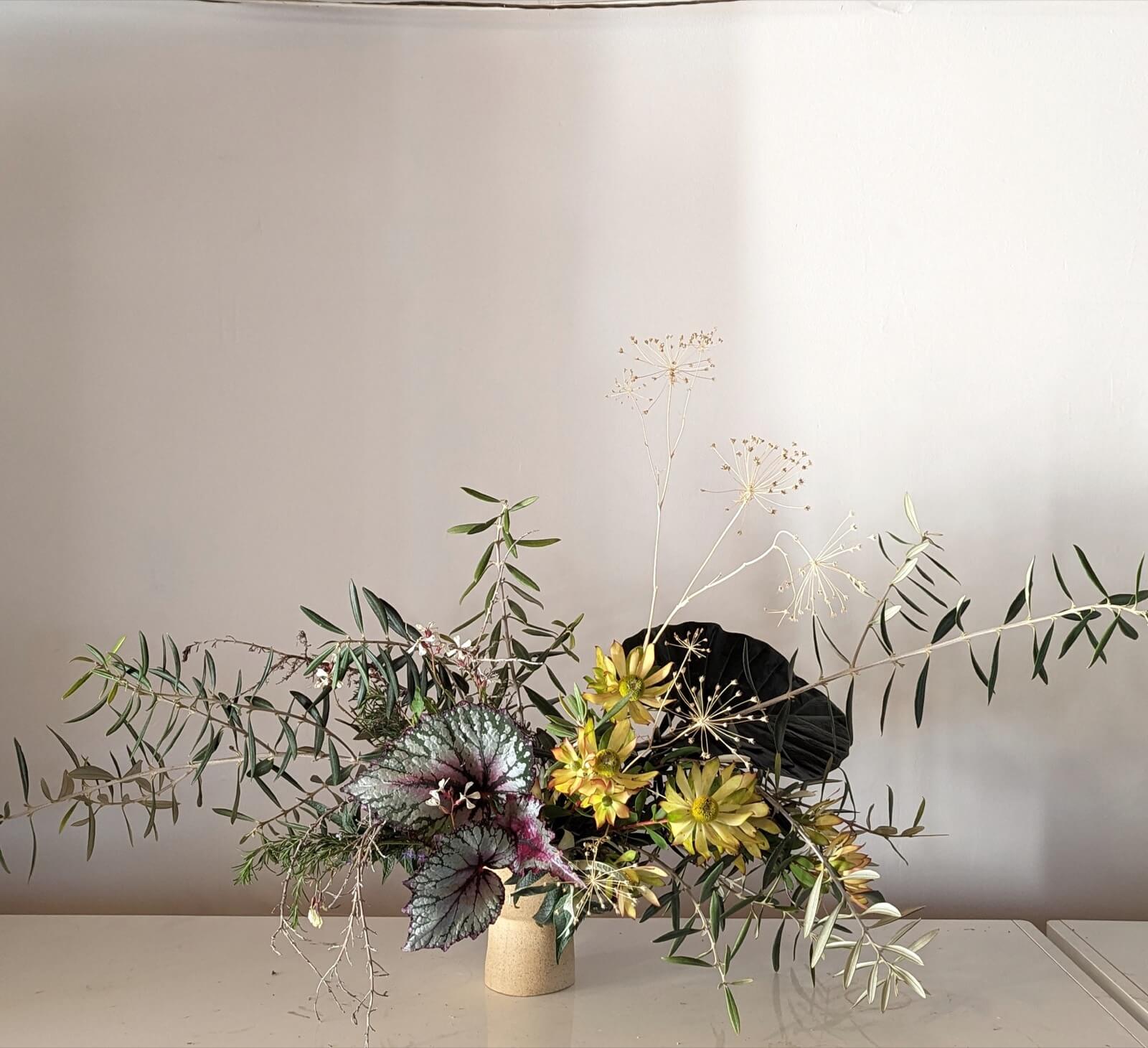 Low sculptural arrangement of olive branches, begonia leaves, and yellow dahlias