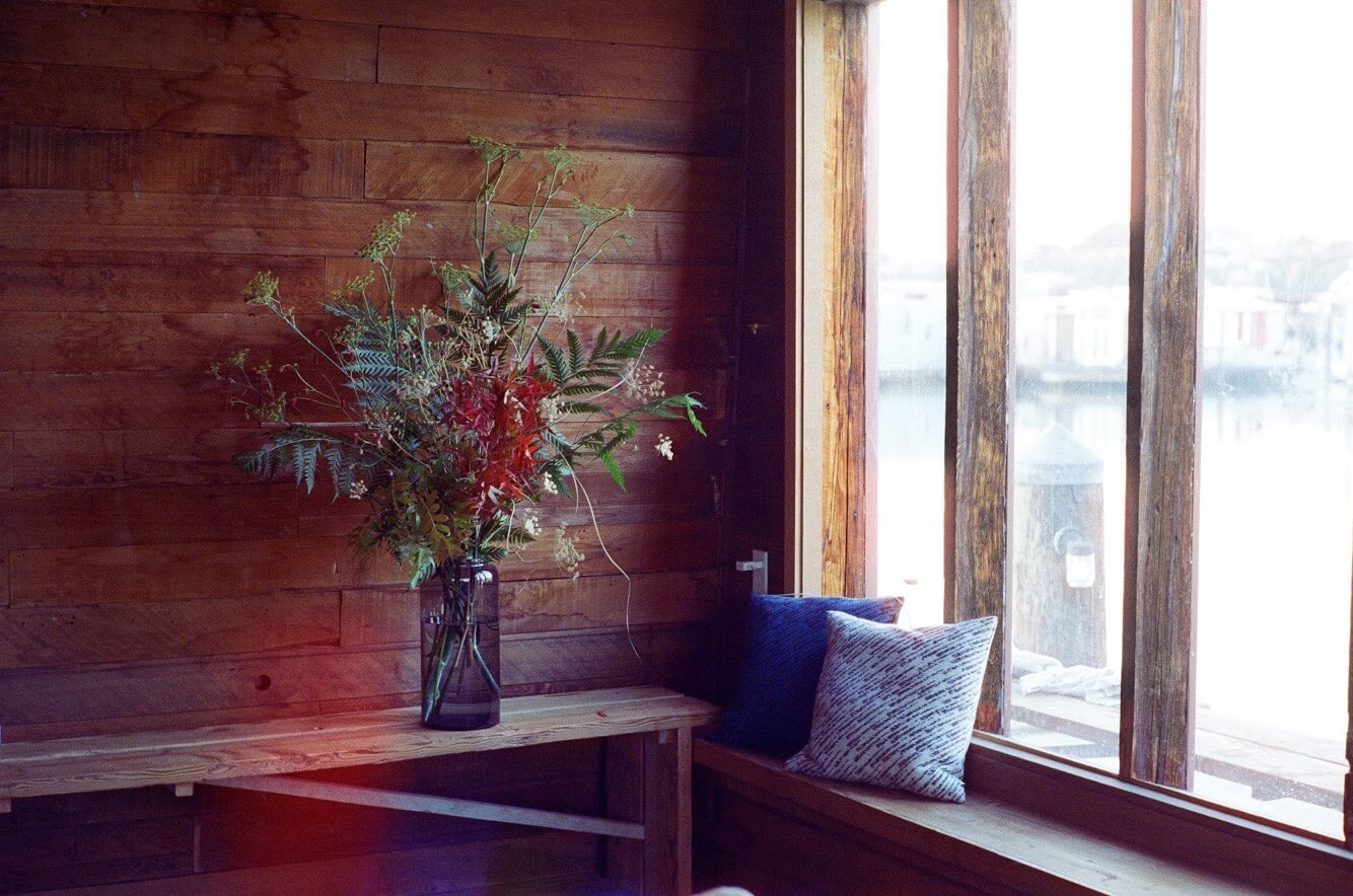 Arrangement of palm and red blooms in a glass vase on a wooden window bench overlooking the water